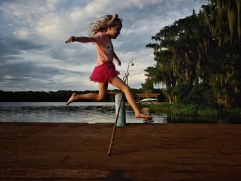 Girl jumping over fishing rod at lakeshore against sky