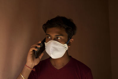 Close-up of young man wearing flu mask talking on phone against wall at home