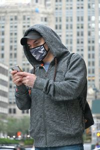 Man wearing mask holding smart phone while standing against building