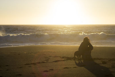 Woman crouching on beach with dog against sky during sunset