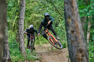 Male cyclists in protective helmets and costumes performing dangerous stunts on bikes for downhill on trail in forest