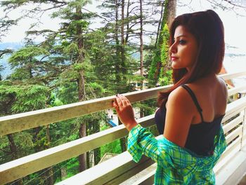 Young woman looking away while standing by railing against trees