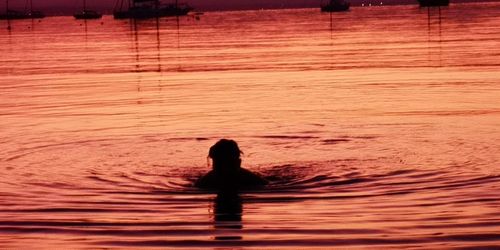 Silhouette person swimming in sea against orange sky