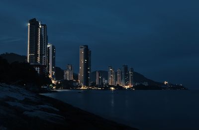 Illuminated city by sea against sky at dusk