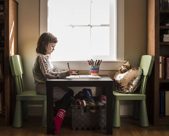 Side view of girl drawing on paper at table against window in home