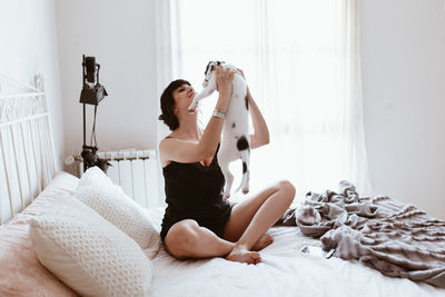 Smiling young woman playing with dog while sitting on bed at home