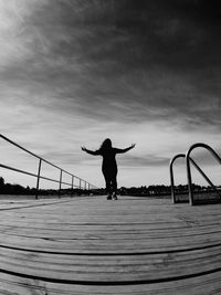 Rear view of silhouette young woman with arms outstretched standing on pier against sky