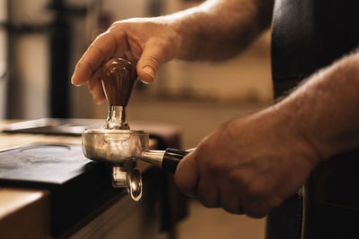 Side view of crop unrecognizable male barista pressing ground coffee with tamper in portafilter while standing at counter with mat during espresso preparation in cafe