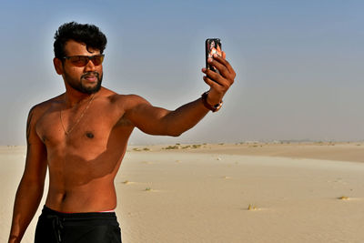 Shirtless young man taking selfie with mobile phone at desert during sunny day