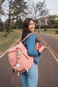 Back view of happy young ethnic female student with backpack and books using mobile phone and looking at camera over shoulder while strolling on asphalt path in green park
