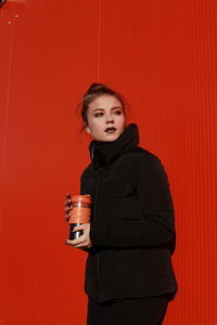 Fashionable teenage girl wearing black warm clothing standing against red wall during sunny day