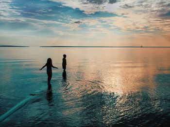 Silhouette girl and boy standing in sea against sky during sunset