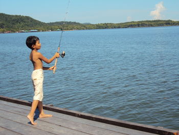 Side view of shirtless boy fishing in lake while standing on pier