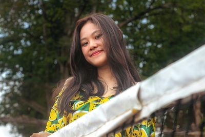 Low angle portrait of young woman standing behind tennis court against trees