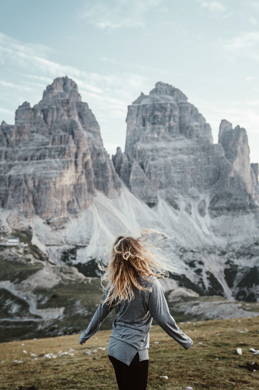 leisure activity, real people, one person, lifestyles, rear view, mountain, beauty in nature, hair, women, scenics - nature, hairstyle, standing, rock, nature, environment, sky, blond hair, rock - object, day, mountain range, outdoors, formation