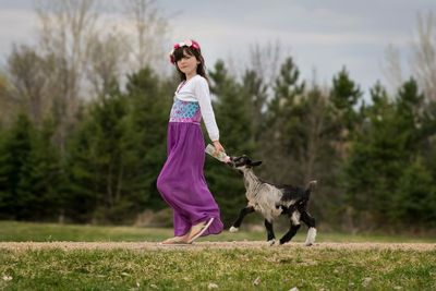 Girl feeding lamb and walking on grassy field against trees