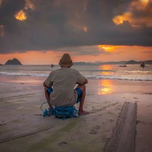 Rear view of man sitting on beach against sky during sunset