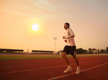 Full length of teenage boy running on sports track against orange sky