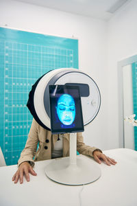 Unrecognizable woman sitting at table in facial skin diagnostics equipment with tablet during procedure in modern beauty clinic