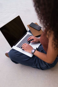 African american self employed woman sitting on street with laptop and browsing smartphone