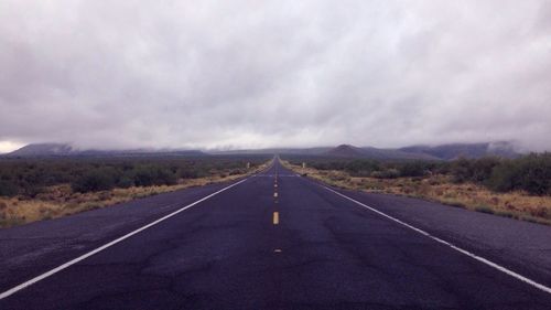 Empty road amidst fields against cloudy sky