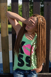 Girl with dreadlocks covering eyes while standing by wooden fence