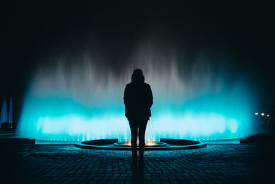 Rear view of silhouette woman standing by illuminated fountain at night