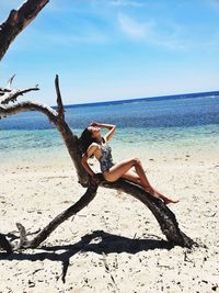 Woman in bikini sitting on tree trunk at beach against sky during sunny day