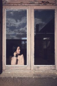 Thoughtful young woman standing by window seen through glass