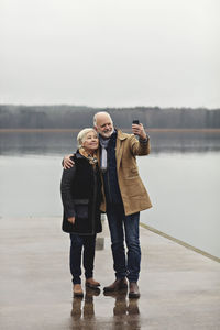 Full length of senior couple taking selfie at lakeside against clear sky