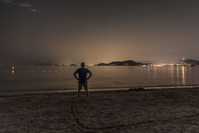 Rear view of man standing at beach against sky at night