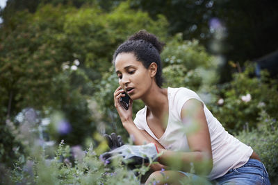 Young female gardener using mobile phone while looking at plants