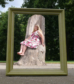 Full length portrait of woman sitting on tree stump seen through picture frame