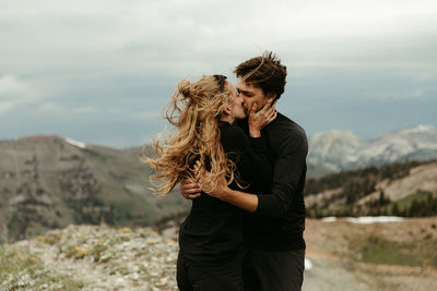 Newly engaged white couple dressed in all black kiss on windy ridge