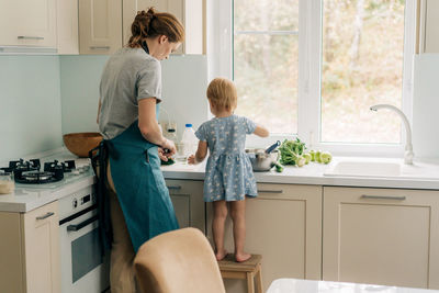 View from the back of a mother with a little daughter cooking in the home kitchen.