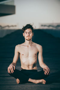 Shirtless young man meditating while sitting on boardwalk
