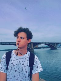 Young man biting beads while standing by river in city against sky