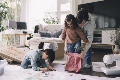 Mother helping daughter in wearing skirt while son drawing on cloth at home