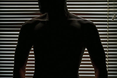 Shirtless silhouette man standing against window blinds