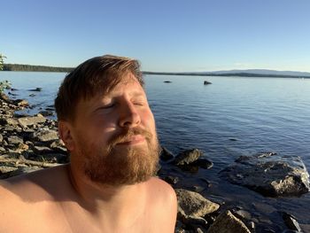 Bearded shirtless man enjoying the sun with closed eyes by a lake.