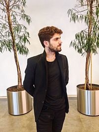 Young man looking away while standing on potted plant against wall