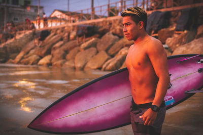 Young shirtless man carrying surfboard at beach