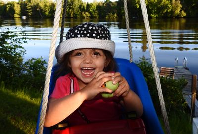 Portrait of smiling cute girl playing on swing against lake