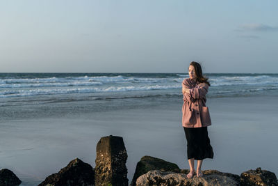 Full length of woman standing on rock at beach against sky