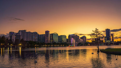 River by illuminated buildings against sky during sunset