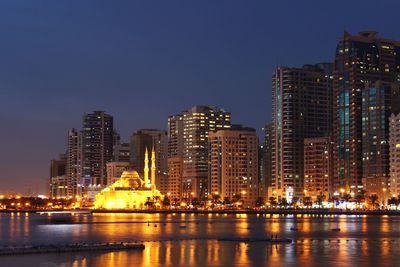 Illuminated mosque by river in city against sky at dusk