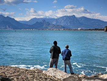 Men fishing while standing on rocky shore against mountains