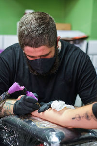 Unrecognizable concentrate bearded male master in black face mask and gloves creating tattoo on arm of crop faceless client in salon