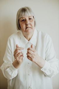 Sad elderly woman in white blouse gesturing with hands on light background looking at camera