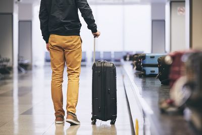 Low section of young man with luggage standing by conveyor belt at airport